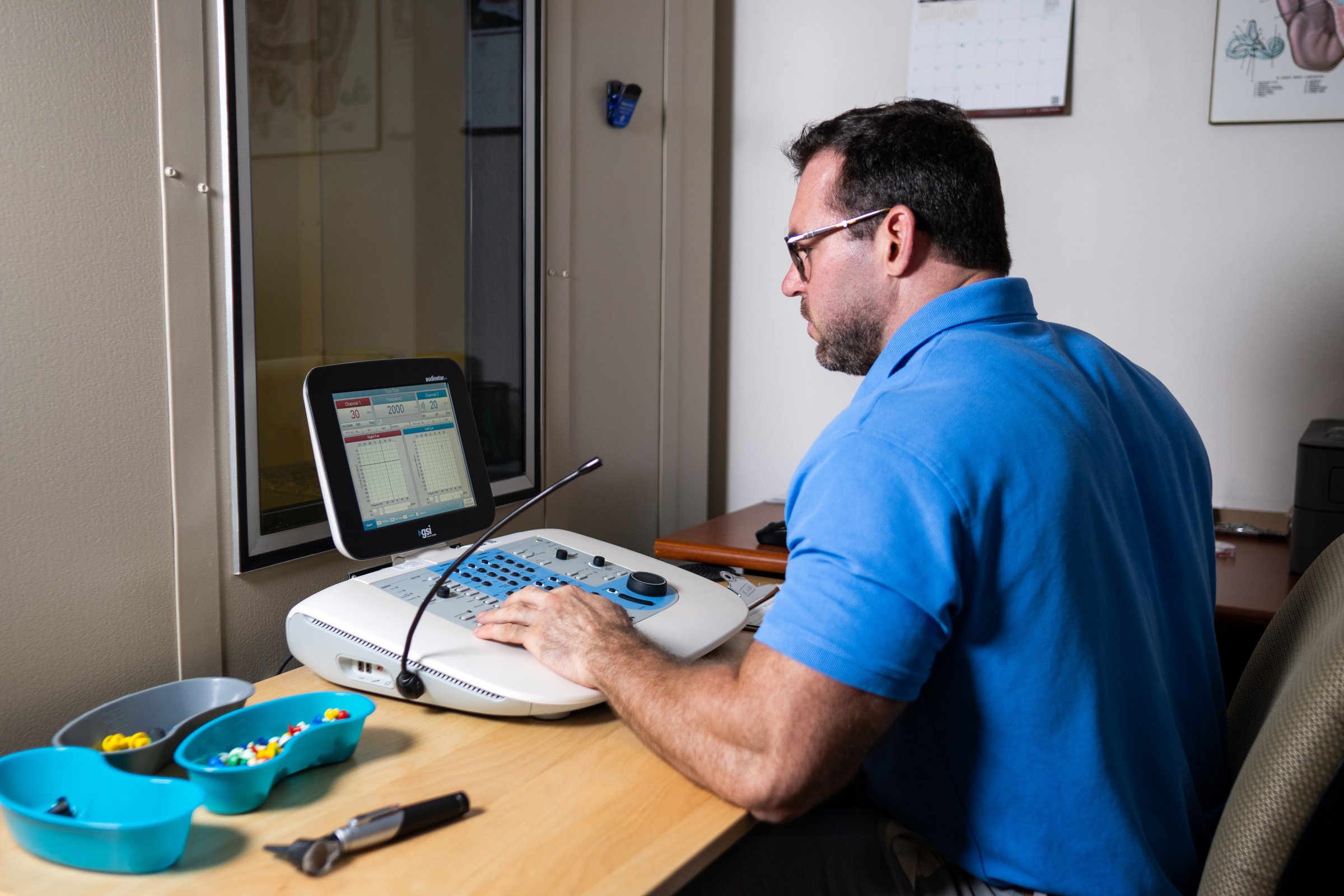Andy Schirmer at the audiometer during a hearing evaluation, with the patient visible in the sound treated booth